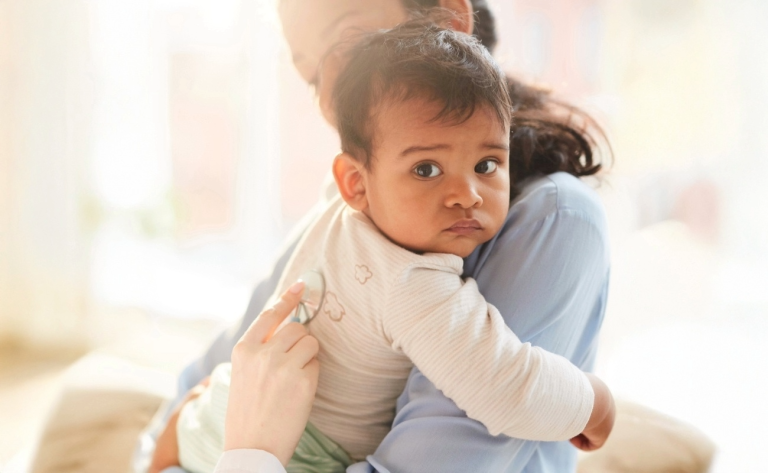 Young mother holding her little child on her hands while female doctor examining him with stethoscope at hospital